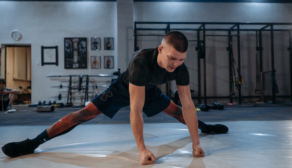 A male athlete stretches intensely in a dimly lit gym environment, preparing for training.